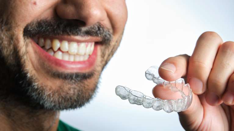 A smiling man holding a clear aligner in his hand, showcasing invisible braces.
